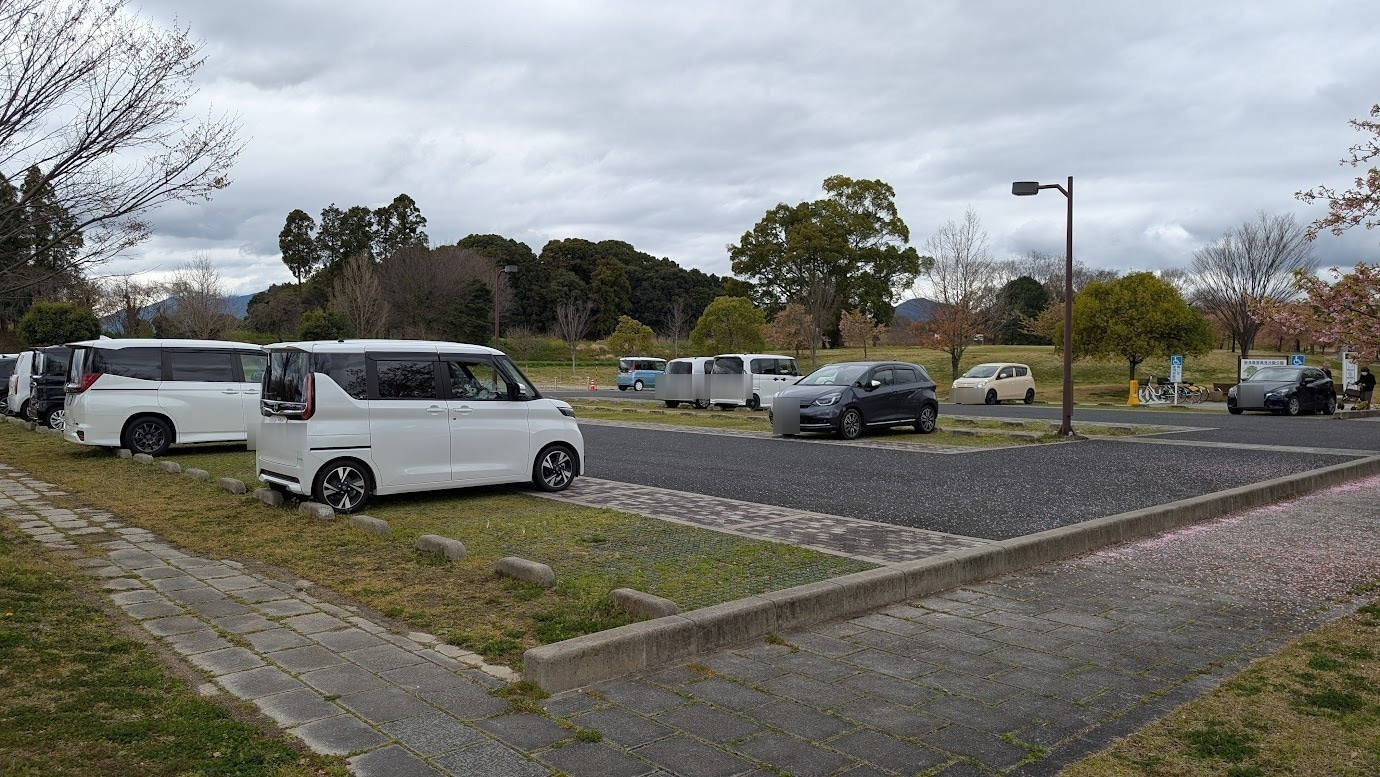 奈良県馬見丘陵公園の南駐車場。平日の昼間、曇り空の下で駐車車両はまばらであり、空きスペースが十分に確保されている。手前には石畳の歩道、奥には豊かな緑の森が広がっている。