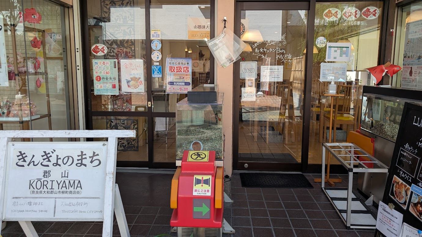 奈良県大和郡山市の「彩食キッチン Bon やなぎ店」の外観。赤いフレームの入口と、手書きで記されたメニューボードが置かれている。