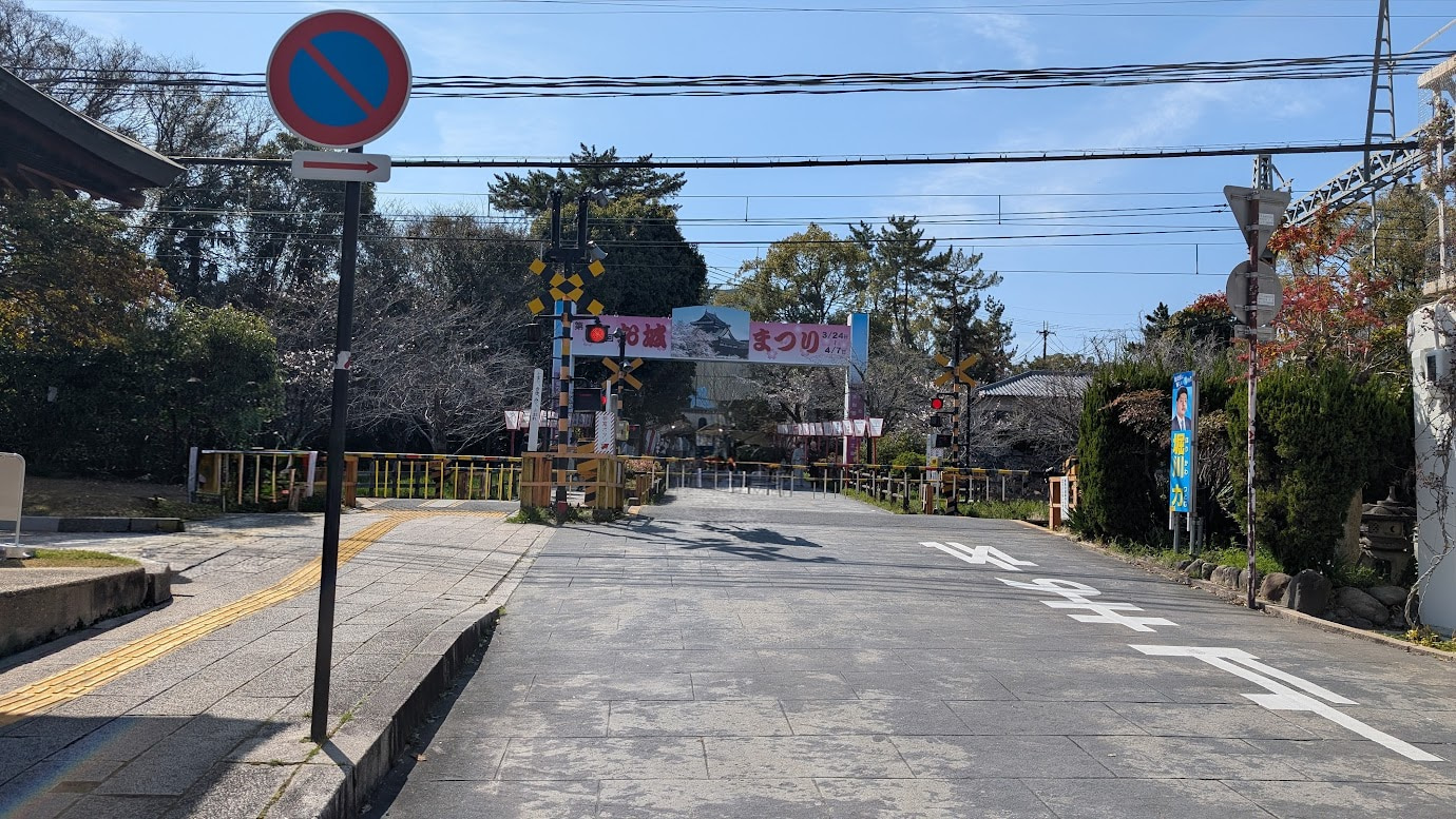 奈良県大和郡山市の郡山城跡で開催されている「お城まつり」の入口。青空を背景に、満開に近い桜と、「第65回 郡山お城まつり」と書かれた大きな看板、多くの提灯が飾られている。