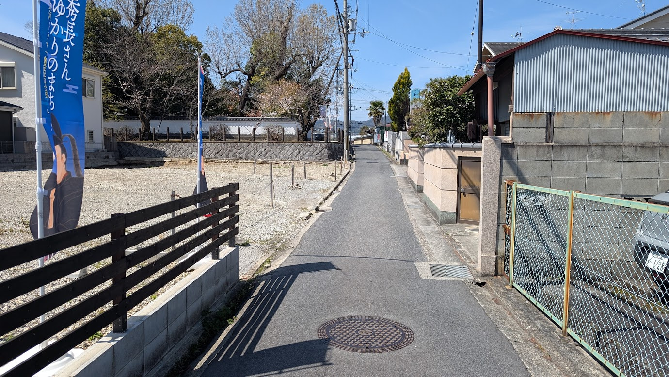 奈良県大和郡山市の大納言塚の入り口へ続く狭い路地。アスファルトの道が住宅の間を通り、奥に大納言塚の石柱と生垣が見える。