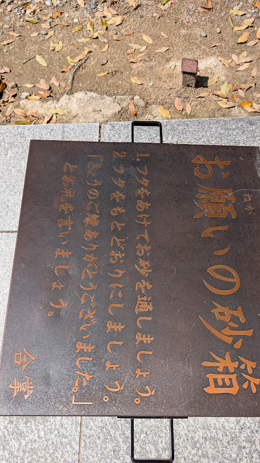 奈良県大和郡山市の大納言塚にある「お願いの砂箱」の案内板。砂の通し方と参拝後の感謝の言葉（合掌）が記されている。