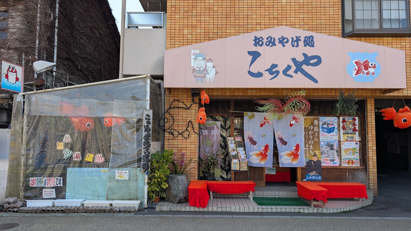 奈良県大和郡山市の「おみやげ処 こちくや」の外観。金魚のイラストが描かれた看板や暖簾、左側には金魚すくい道場の入り口が見える。