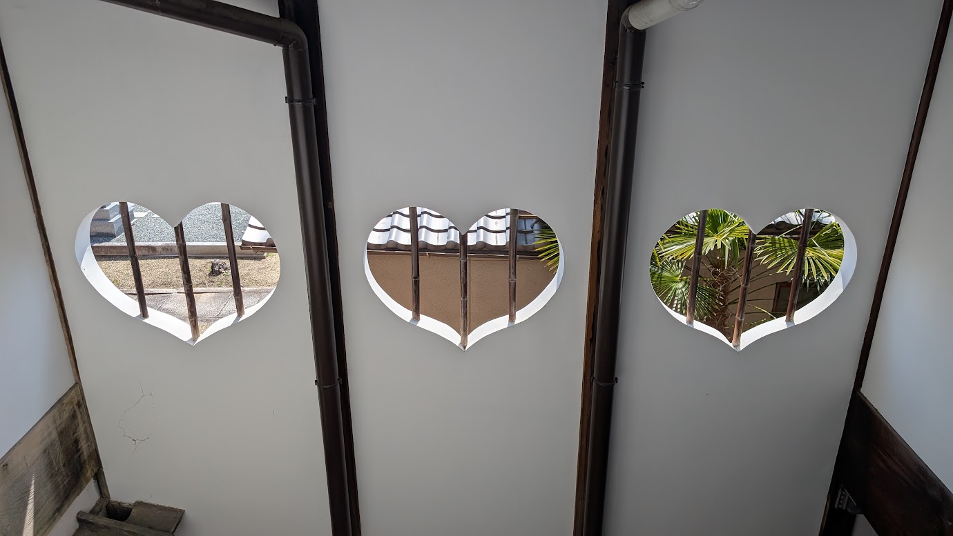 奈良県大和郡山市の町家物語館にある、3つのハート型（猪目型）の窓。白い壁から外の中庭や屋根、木々が覗いており、レトロで美しい光景。