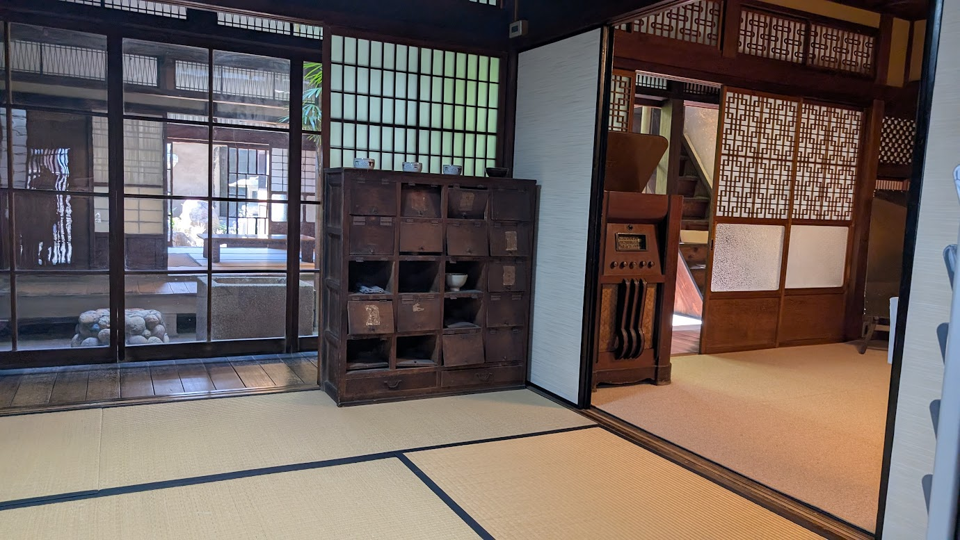 奈良県大和郡山市の町家物語館（旧遊郭建築）の内部。手前には畳敷きの和室があり、奥には中庭の緑が見える。古い木製の棚や格子戸がレトロな雰囲気を醸し出している。