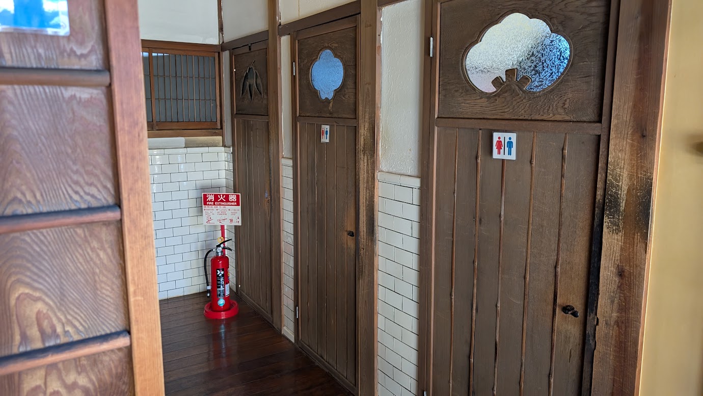 奈良県大和郡山市の町家物語館にあるお手洗い。レトロな木製扉と白いタイルが美しく、消火器が設置された清潔感のある廊下。男女別のマークも完備されている。