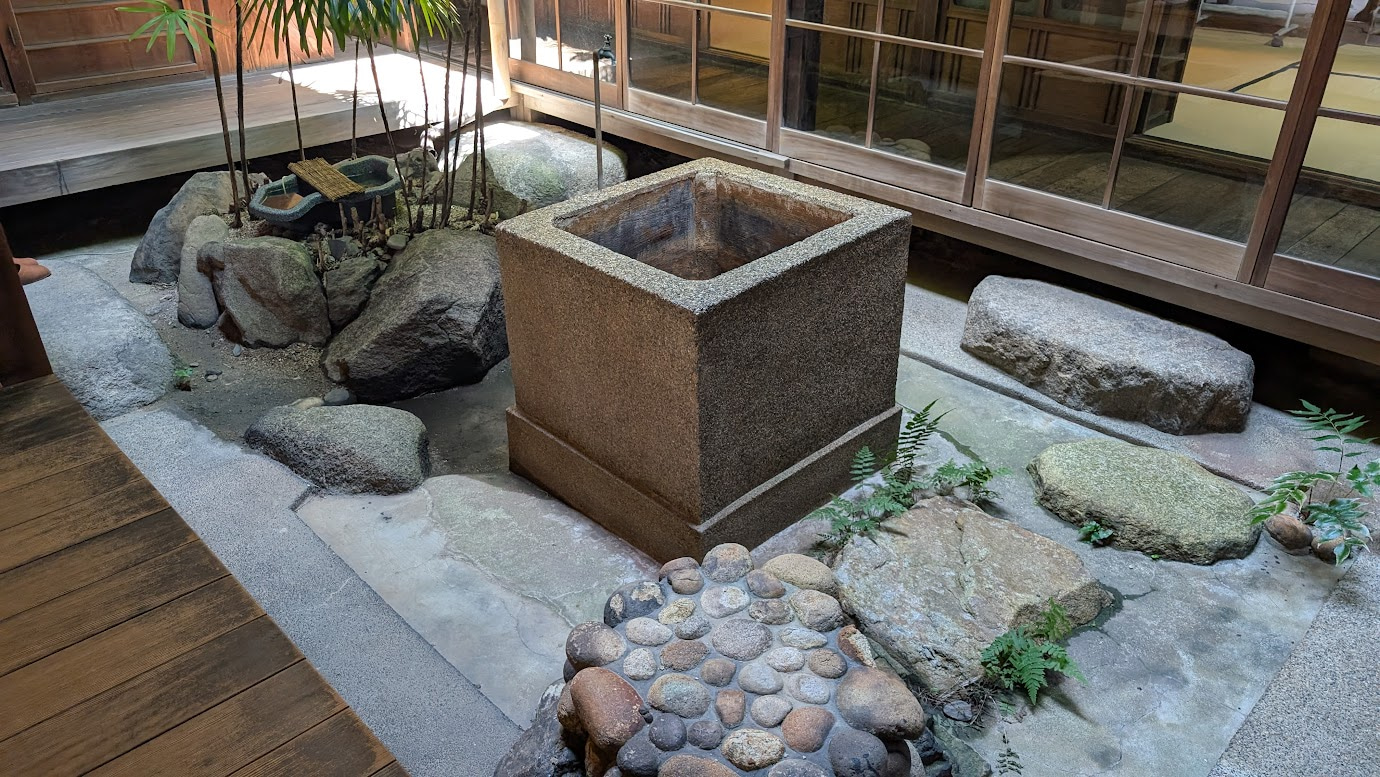 奈良県大和郡山市の町家物語館（旧遊郭建築）にある中庭。中央に四角い石造りの水受けがあり、周囲には自然石や緑のシダ植物が配置され、建物の縁側に囲まれている。