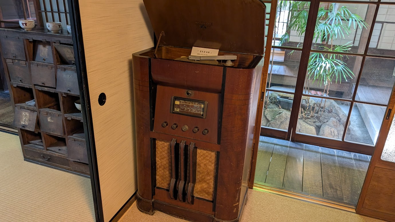 奈良県大和郡山市の町家物語館にある、昭和22年製の電気蓄音機。重厚な木製の筐体で、横には使い込まれた古い多引き出しの箪笥があり、奥には中庭の緑が見える。