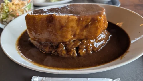 明日香村にある「工場内食堂 ぶたと〇〇と。」の看板メニュー「超ブタカレー」のアップ写真。ご飯の上に信じられないほど分厚く巨大な豚肉の塊が乗っており、濃厚なカレールーがたっぷりとかかっているインパクト抜群の風景。