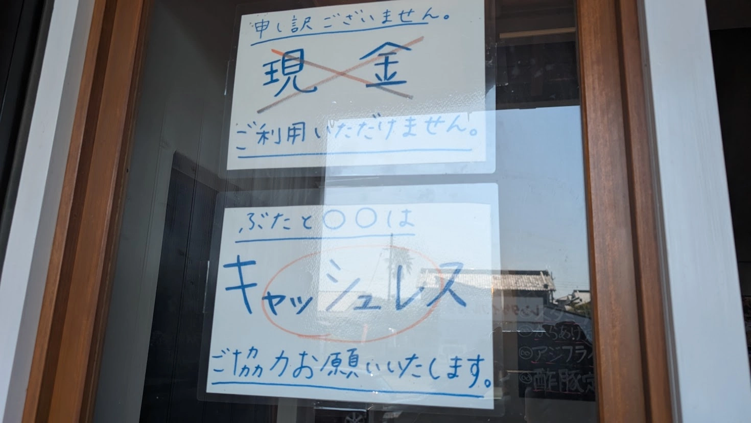「工場内食堂 ぶたと〇〇と。」の入り口ドアに貼付された手書きの案内書き。「現金ご利用いただけません」「完全キャッシュレスご協力お願いいたします」と青い文字で注意喚起されている風景。