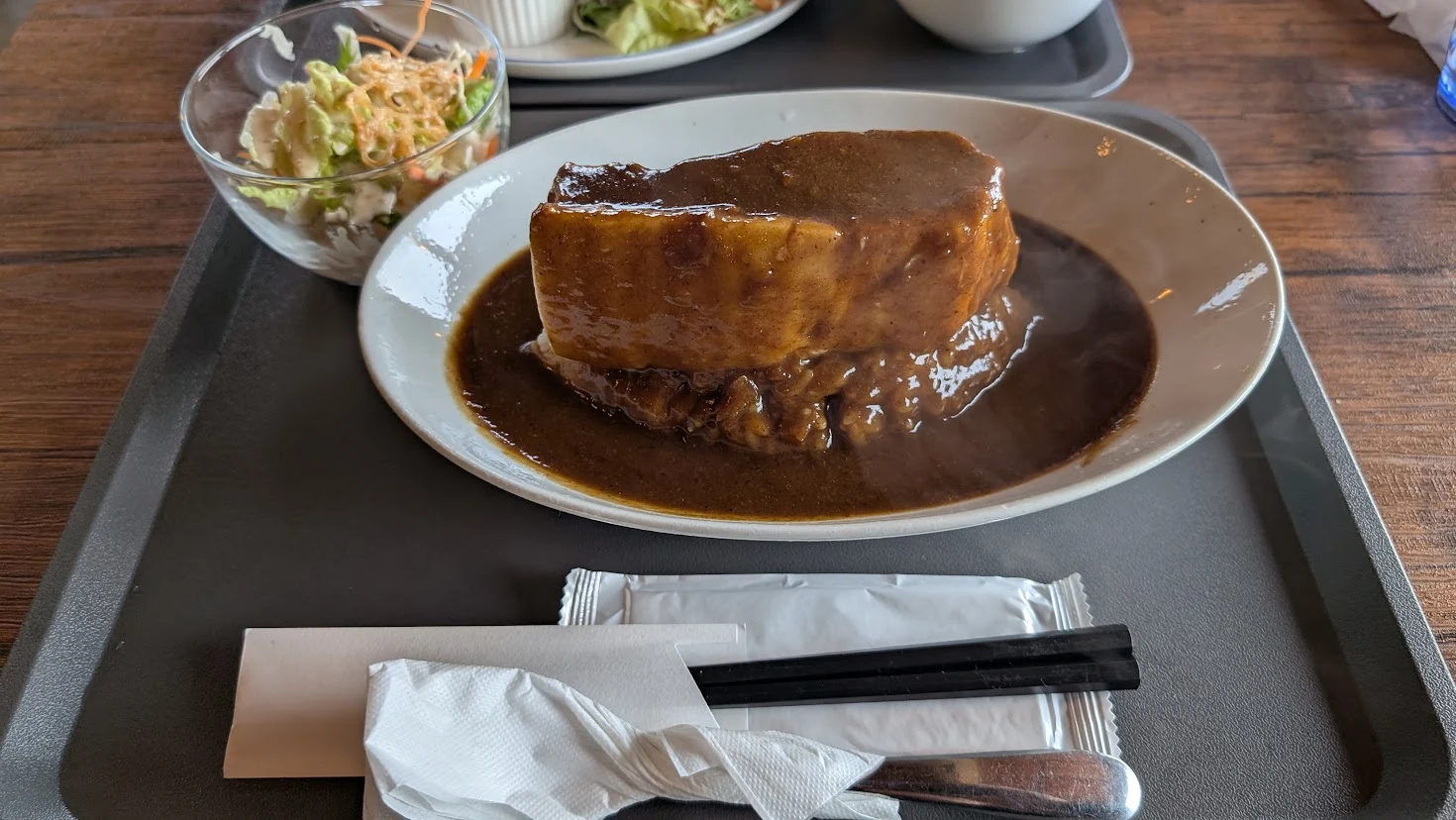 明日香村の「工場内食堂 ぶたと〇〇と。」で提供される「超ブタカレー」の引きの写真。お盆の上に、巨大な豚肉が乗ったカレーライスと、セットのサラダ、お箸などが並んでいる様子。