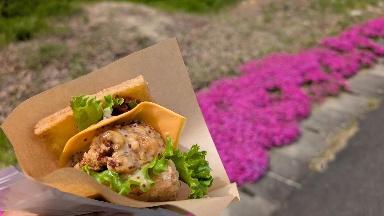 芝桜のピンクを背景に、手で持った厚揚げバーガーのアップ。カリッと焼かれた厚揚げの間に、ジューシーなチキン、とろけるチーズ、レタス、ソースがたっぷり挟まっている