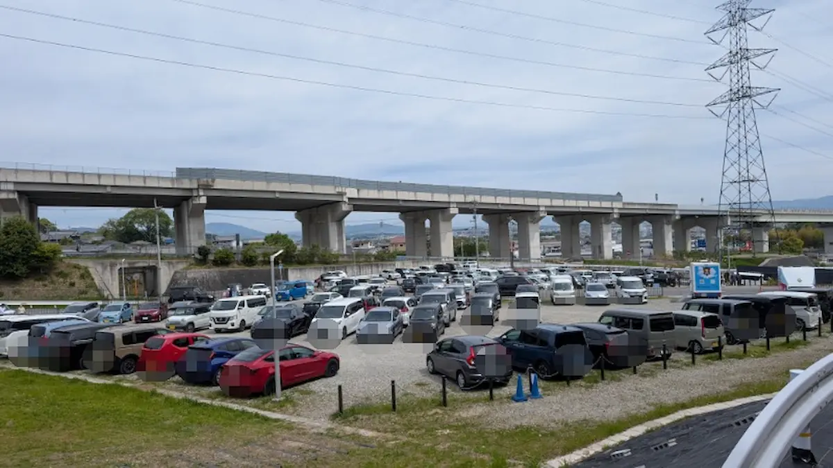 葛城市の芝桜まつり臨時駐車場。午前10時半時点で砂利の駐車スペースがほぼ全ての車で埋まり、混雑している様子。背景には高架道路が続いている