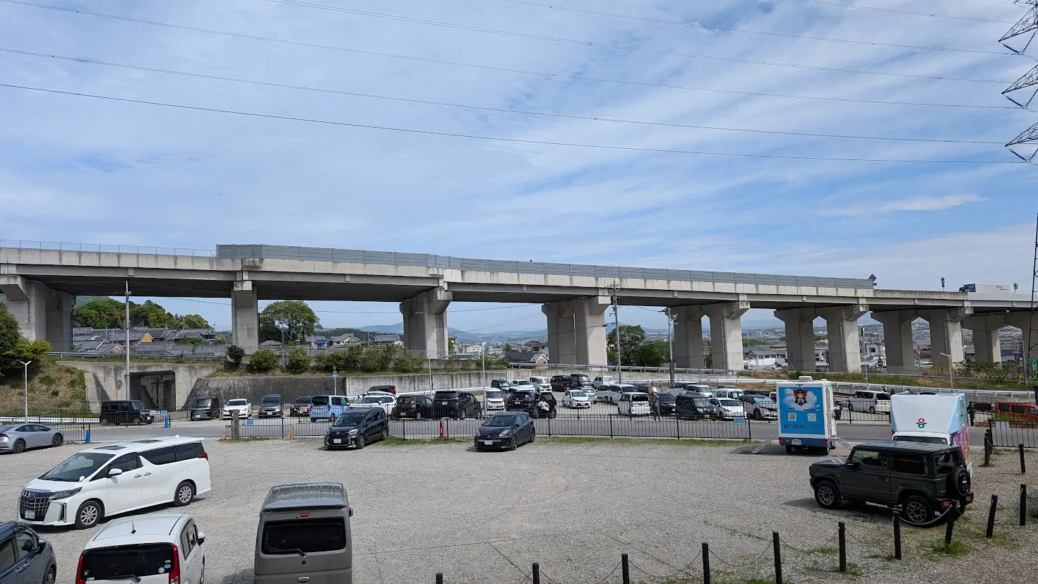 葛城市の芝桜まつり会場付近にある、砂利が敷かれた未舗装の広い駐車場。周囲には緑の山々と、駐車区画を整理するコーンが並んでいる風景