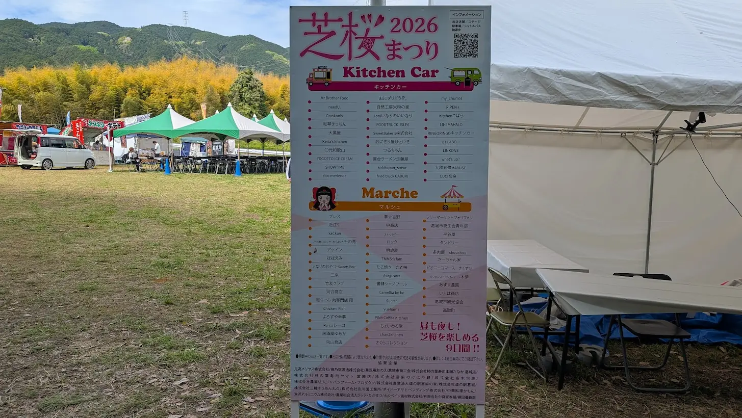 葛城市芝桜まつりの会場に設置された案内看板。キッチンカーやマルシェの出店名が多数並び、背後にはイベントテントと新緑の山が見える風景