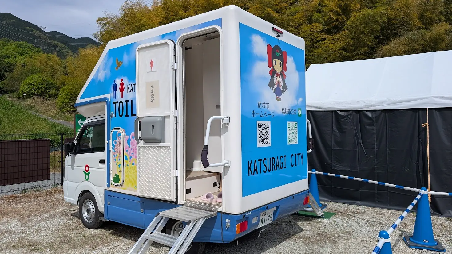 葛城市の芝桜まつり会場の散策路脇に停車している、青い空と雲、蓮花ちゃんがデザインされた軽トラック型の移動式トイレ。入口にはスリッパが置かれている