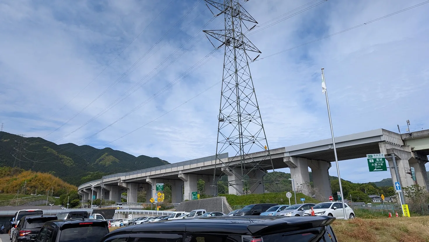 葛城市の道の駅「かつらぎ」第1駐車場。入口付近まで車が連なり、混雑している様子。背景には高架道路と葛城の山々が見える