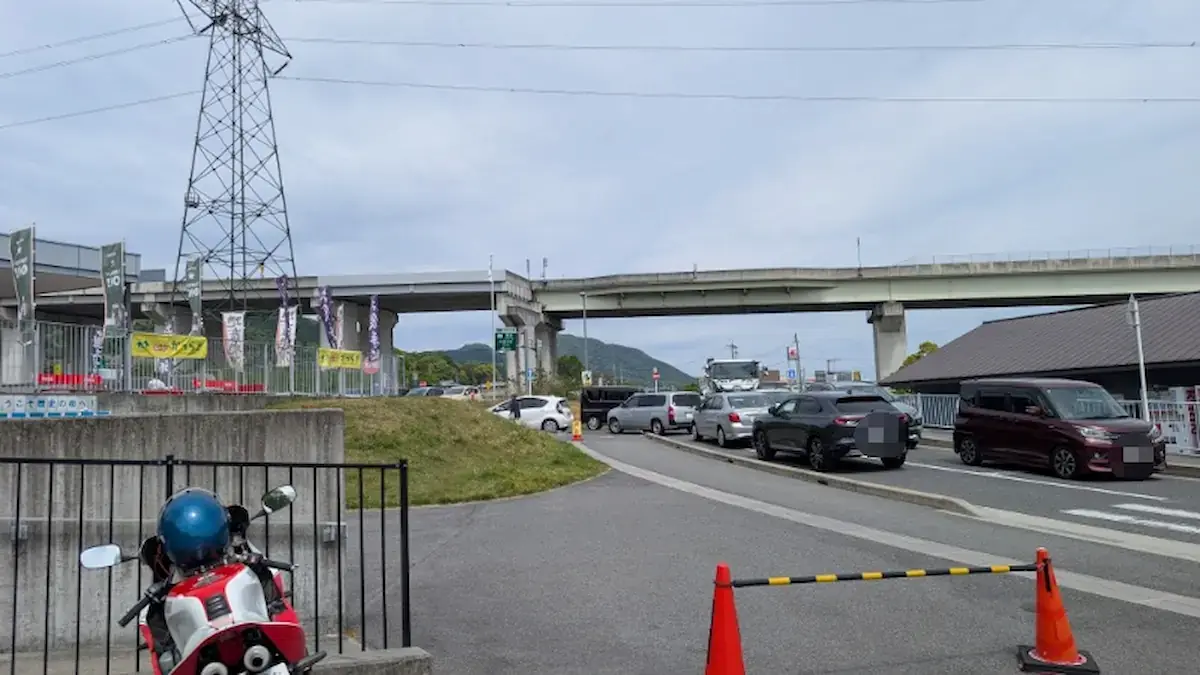 葛城市の道の駅「かつらぎ」前。午前10時半時点で、駐車場へ入ろうとする車が道路に列を作り、混雑している様子。手前にはオートバイ、背景には高架道路が見える