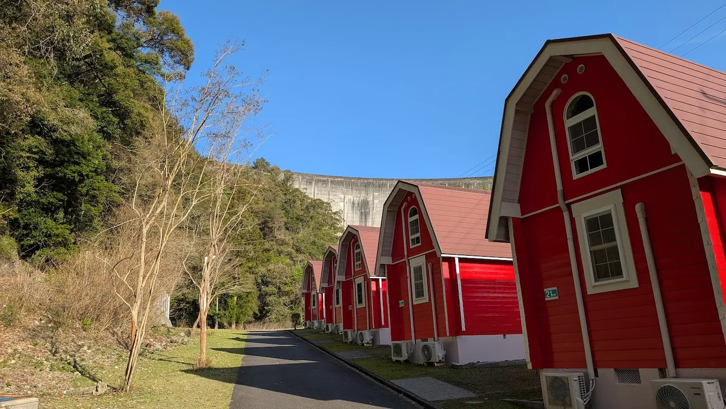 奈良県下北山村「きなりの郷」にある赤い屋根と壁のオートロッジが並ぶ風景。背後には池原ダムの巨大なコンクリート壁面と、新緑の山々が広がっている