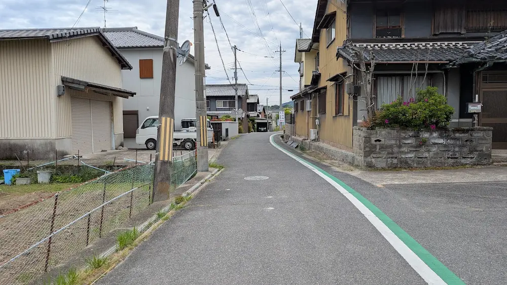 奈良・石光寺へ続く門前の生活道路。普通車一台が通れる程度の道幅で、右側には住宅、左側には電柱や畑がある。路側帯には緑の線が引かれている風景