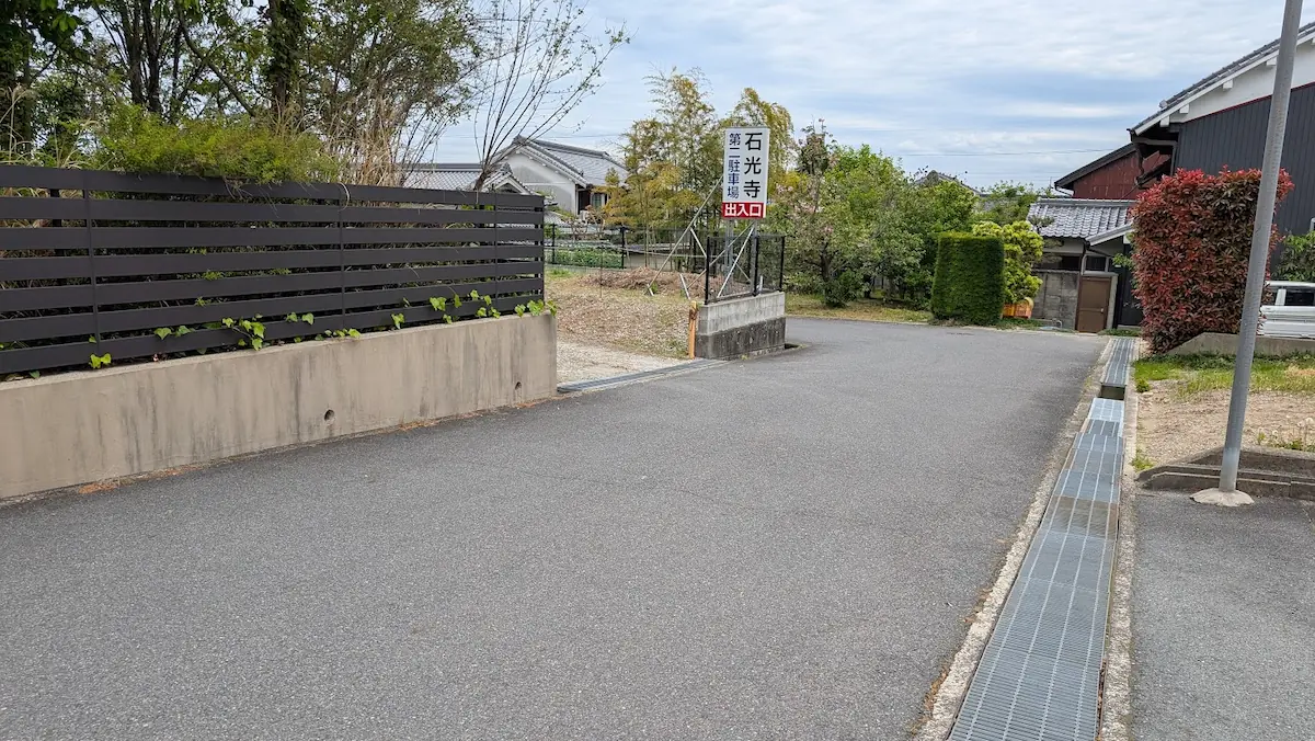 奈良・石光寺の第2駐車場入り口。アスファルトの道路の先に「石光寺 第二駐車場 出入口」と書かれた白い看板が立っており、入り口付近の道幅が少し狭くなっている風景