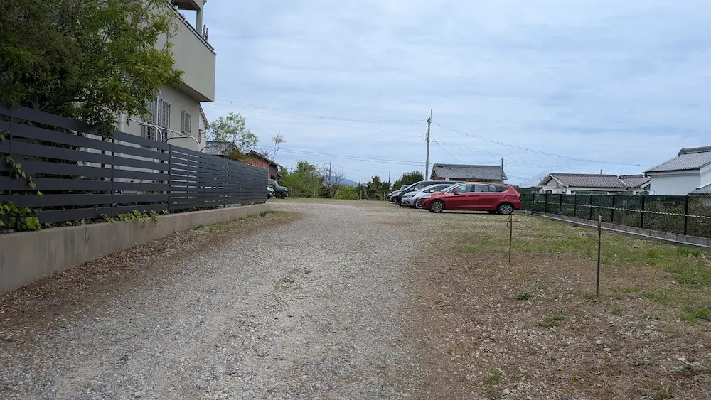 奈良・石光寺の第2駐車場内部。砂利敷きの広大なスペースに車が数台停まっており、周囲には住宅やフェンスがある開放的な駐車場の風景