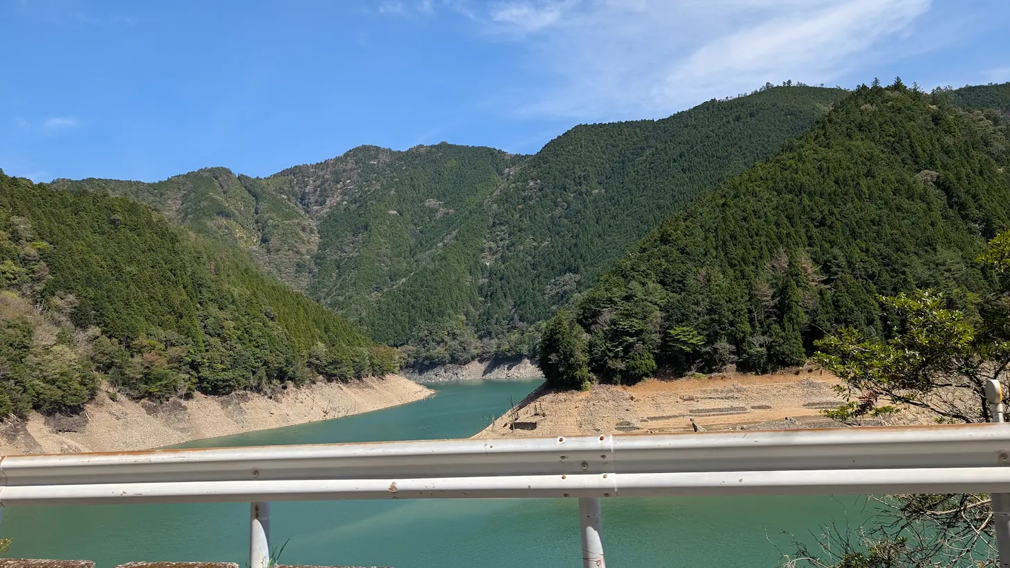 国道のガードレール越しに望む、池原貯水池の風景。深い緑に包まれた山々の間を、静かなエメラルドグリーンの水面が蛇行しながら続いている
