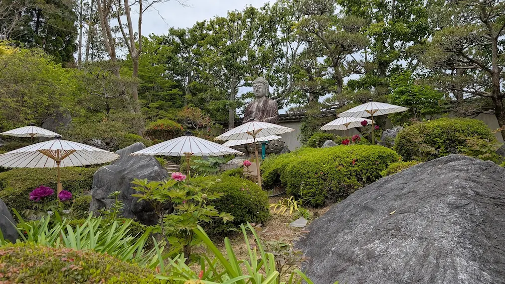 奈良・當麻寺奥院の浄土庭園にて、大きな石組みの間に佇む阿弥陀如来像。手前には和傘に守られた鮮やかなピンクの牡丹が咲き、新緑の木々に囲まれている風景