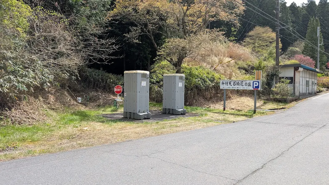 宇陀・弁財天の駐車場入口付近に設置された2基のグレーの仮設トイレ。横には「弁財天 石楠花の丘 P」の看板と手洗い用の蛇口が見える風景