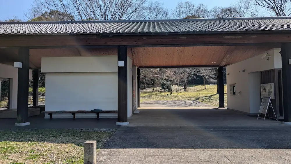 3月の甘樫丘にある北側休憩所の外観。茶色い瓦屋根とベージュの壁で造られたログハウス風の平屋建ての建物で、入り口には自動販売機やトイレの案内、観光情報が掲示されている。