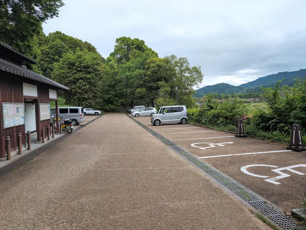甘樫丘の麓にある「川原駐車場」のドローン空撮写真。平日午前9時頃の様子で、広い駐車場に数台の車が停まっている。周囲は新緑の木々に囲まれ、奥には明日香村の田園風景が広がっている。