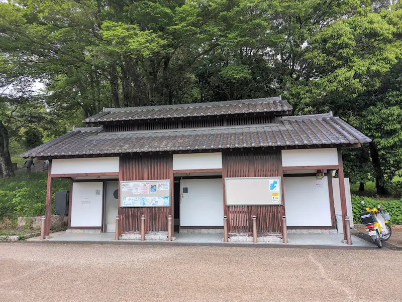 甘樫丘・川原駐車場の公衆トイレ。飛鳥の景観に配慮した瓦屋根と木目調の外壁が特徴的な和風建築。左側には多目的トイレ、中央には掲示板があり、右端には黄色いバイクが停まっている。