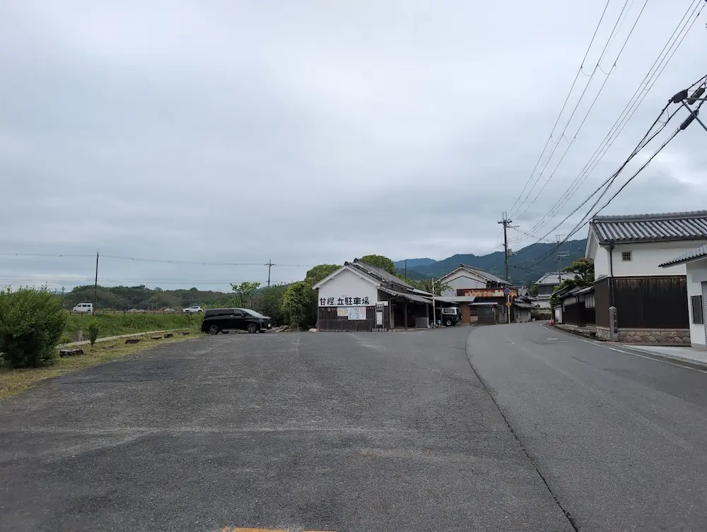 甘樫丘の麓、県道沿いにある民間の有料駐車場。白い壁に「甘樫丘駐車場」と大きく書かれた建物が目印。手前は舗装されており、道一本挟んで砂利の駐車場と隣接している。