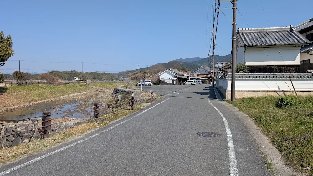甘樫丘北側休憩所のすぐ近くから明日香夢の楽市へと続く、広くて舗装された見通しの良い道路。右側には趣のある白壁の民家、左側には小川(飛鳥川)が流れ、その向こうにはなだらかな山々が連なっている。