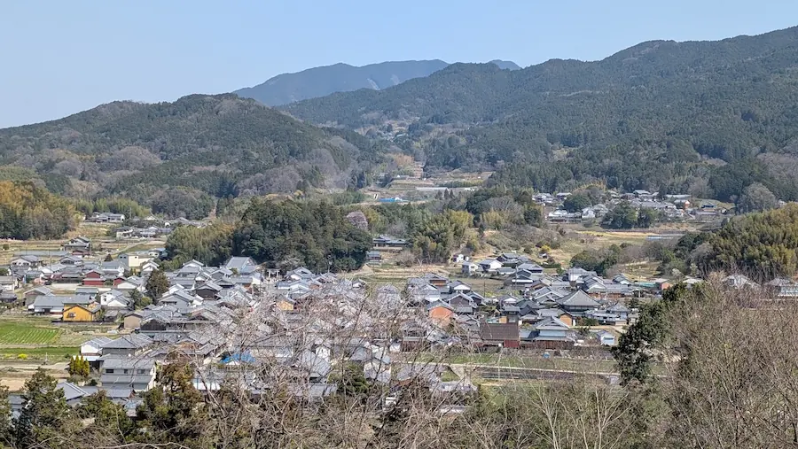 3月の甘樫丘展望台から見渡す明日香村の集落。冬枯れの木々の向こうに、伝統的な瓦屋根が並ぶ家々と、段々畑、そして幾重にも重なる大和の山々が、澄んだ青空の下でくっきりと写っている。