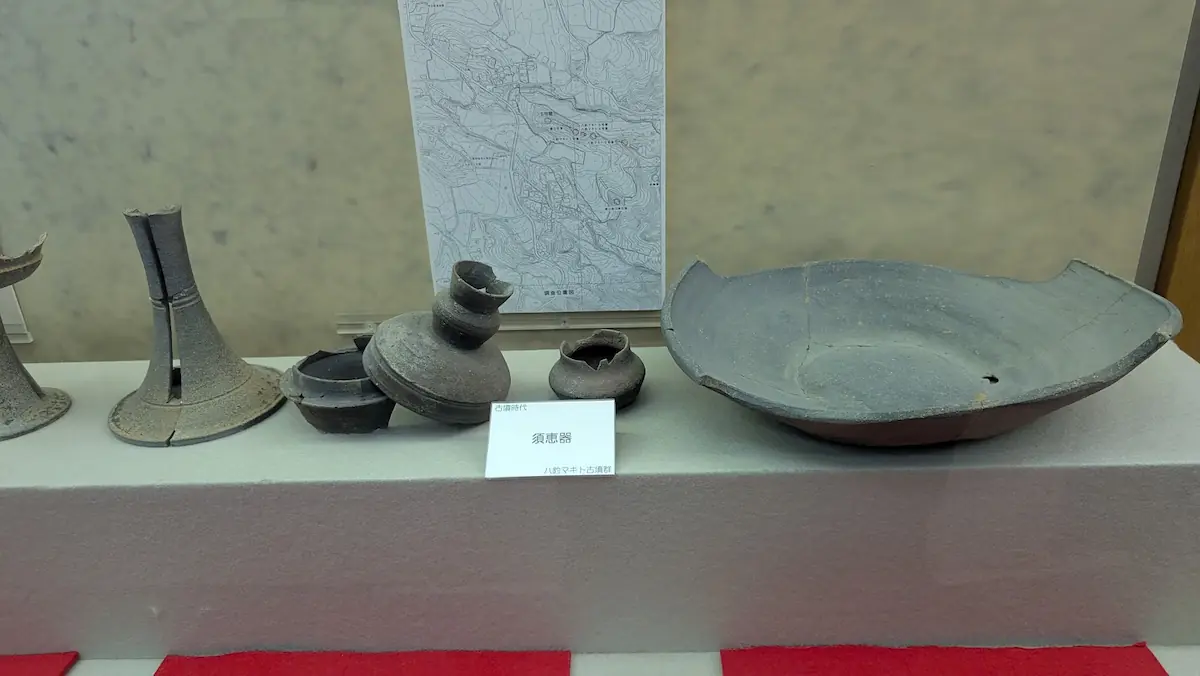 明日香村埋蔵文化財展示室の展示。古墳時代に作られた須恵器が並んでおり、左から縦長の脚台、蓋付きの小鉢、平たい大きな皿状の器などが展示されている。中央には「須恵器 八釣マキト・古墳群」と書かれた札がある。