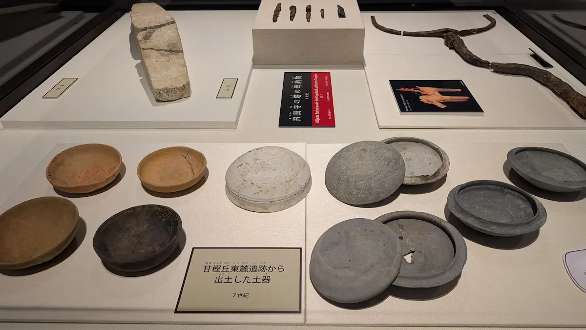 飛鳥資料館の展示ケース。手前には甘樫丘東麓遺跡から出土した7世紀の土器(皿や蓋付きの器)が並び、奥には飛鳥寺の塔の埋納物などが展示されている。