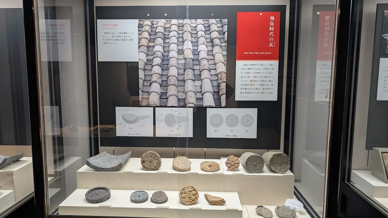 飛鳥資料館の瓦展示。上段には元興寺の屋根写真、下段には飛鳥寺などから出土した蓮華文の軒丸瓦が並んでいる。図解パネルでは瓦の構造や文様の変遷が説明されている。