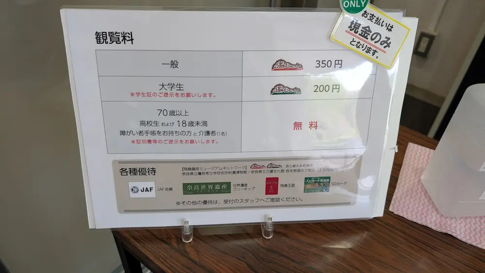 飛鳥資料館の入口看板。石人像のレリーフが施された銀色の看板の奥に広い駐車場がある。2枚目は観覧料金表。一般350円、大学生200円、高校生以下無料。