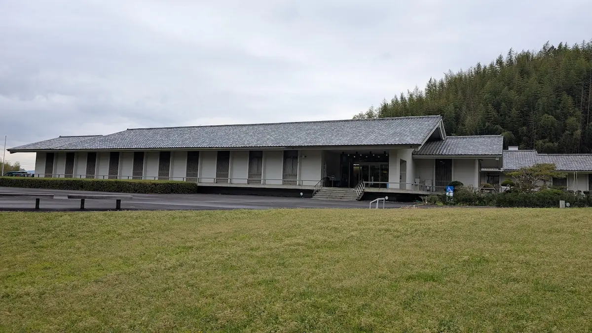 奈良県明日香村にある「飛鳥資料館」の外観全体。瓦屋根の平屋建築が、手前の広い芝生と背後の竹林に囲まれて静かに佇んでいる。