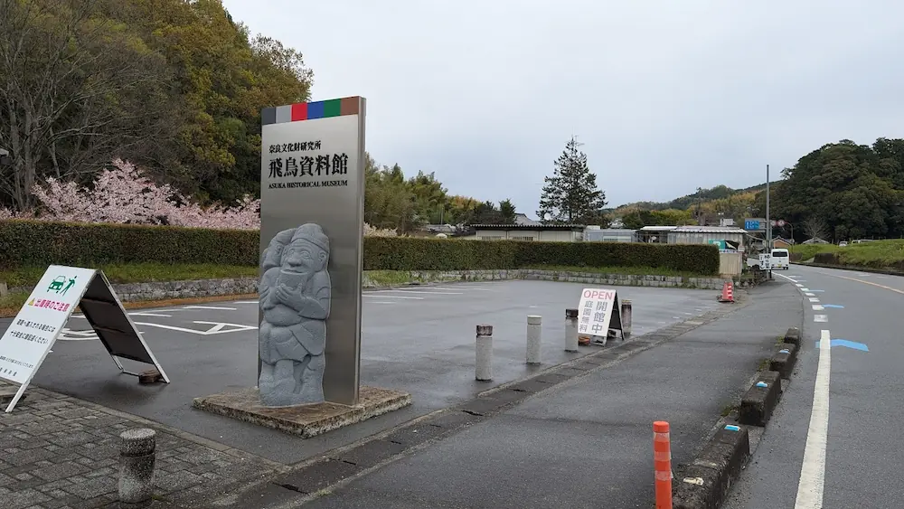 飛鳥資料館の無料駐車場。舗装された広い駐車スペースが広がり、手前には「飛鳥資料館駐車場」の看板と、開場・閉場時間の案内板が立っている。