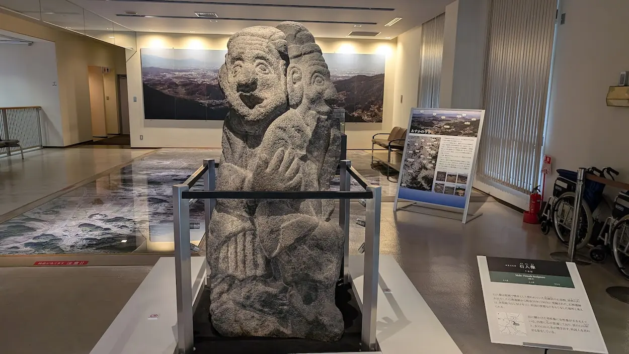 飛鳥資料館のエントランスホールに展示されている「石人像」のレプリカ。男女が背中合わせに寄り添うようなユーモラスな表情の石造物で、手前には解説パネル、奥には床一面の航空写真マップがある。