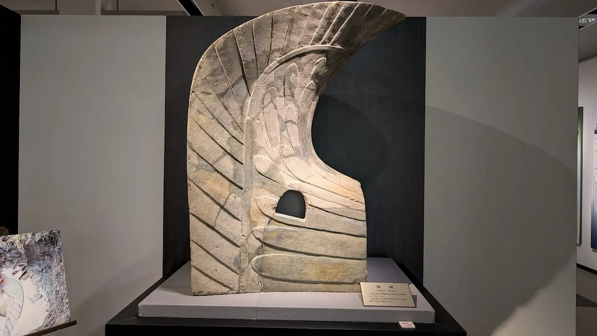 飛鳥資料館に展示されている巨大な鴟尾。鳥の翼のような曲線美を持つ陶器製の屋根装飾で、暗い背景にスポットライトで照らされている。