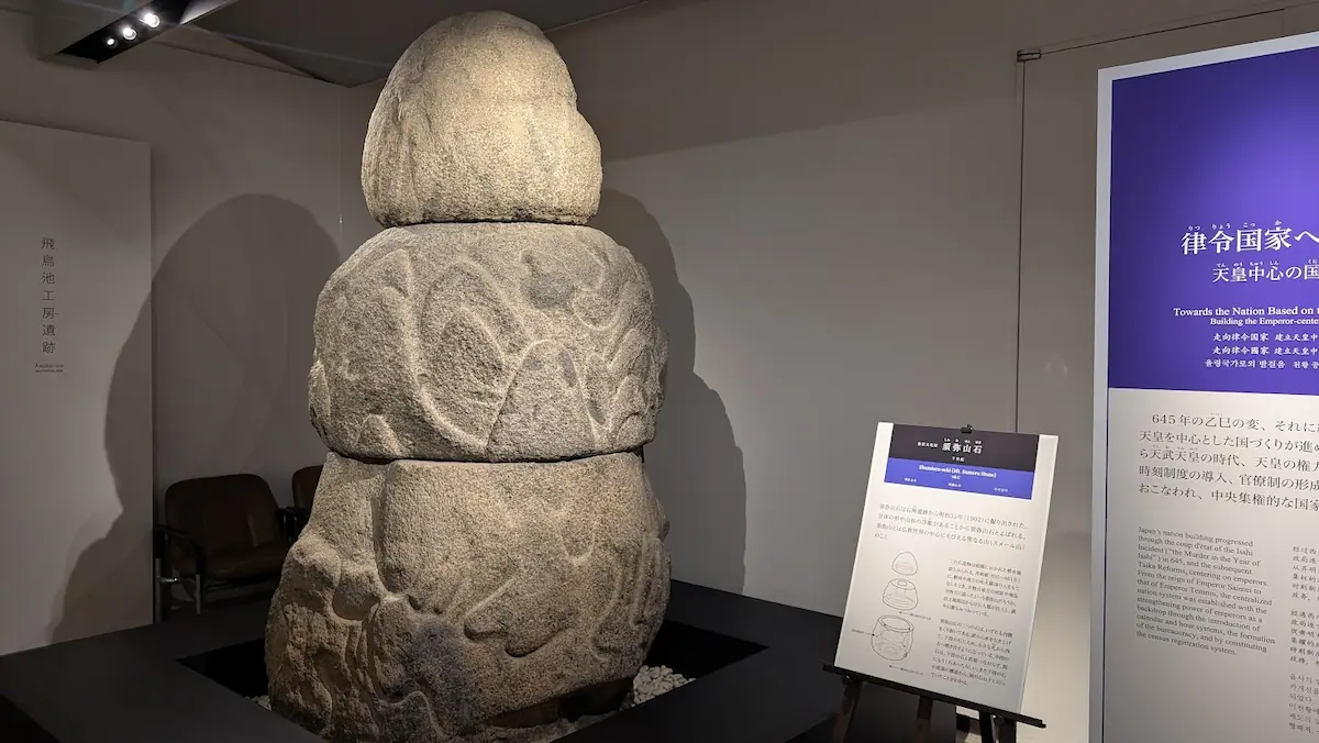 飛鳥資料館に展示されている須弥山石(レプリカ)。3段に積み上げられた巨大な花崗岩に、山岳のような文様が彫り込まれている。右側には解説パネルがある。