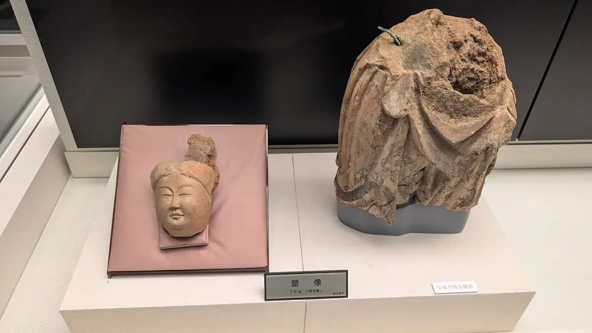 飛鳥資料館に展示されている7世紀の塑像。左側には優しく微笑む仏像の顔、右側には緻密な衣の文様が残る胴体部分の断片が並んでいる。