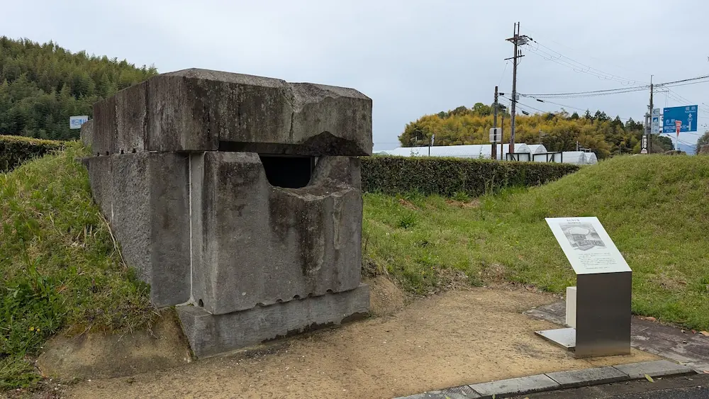 飛鳥資料館の庭園にある高松塚古墳石室のレプリカ。精巧に切り出された巨大な石組みの模型で、横には解説パネルがあり、背景には電柱や標識が見える。