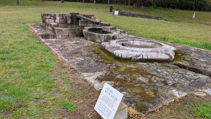 飛鳥資料館の庭園にある亀形石造物の復元展示。亀の形をした石の槽に水が流れ込む様子が再現されており、手前には解説パネル、背景には芝生が広がっている。
