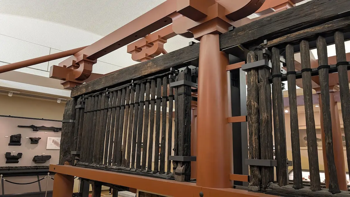 山田寺東回廊のアップ。土の中から掘り出された黒い古材の連子窓(れんじまど)と、それを支える現代の赤い木組み。古材の表面には縦に走る木目や欠けがリアルに残っている。