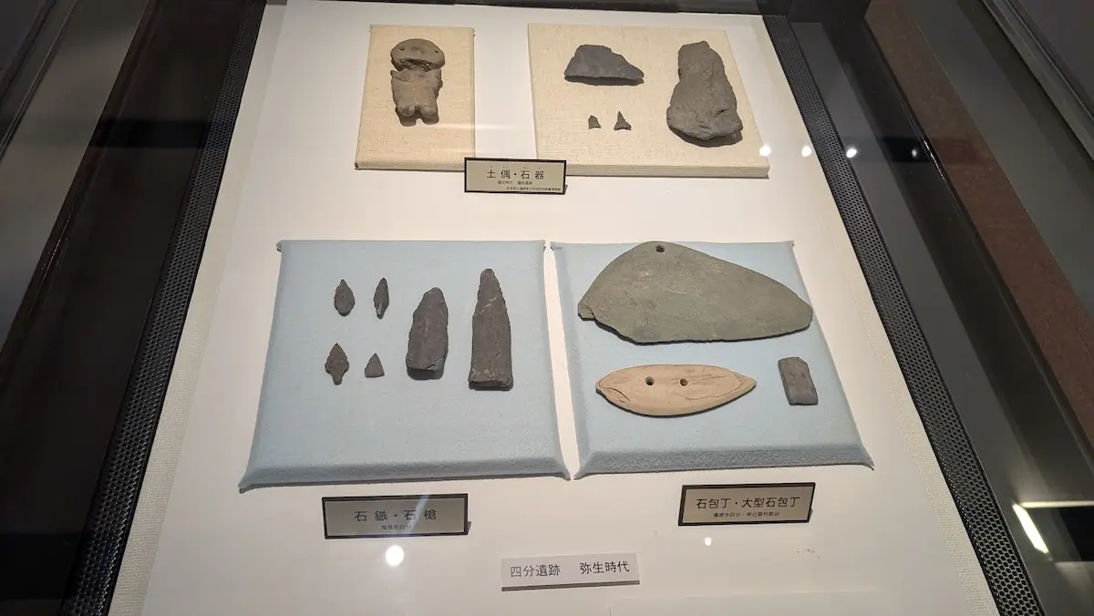 飛鳥資料館に展示されている、弥生時代(四分遺跡)の石器類。石鏃(いしぞじ)、石槍、石包丁、大型石包丁などが丁寧に並べられ、解説プレートが添えられている。