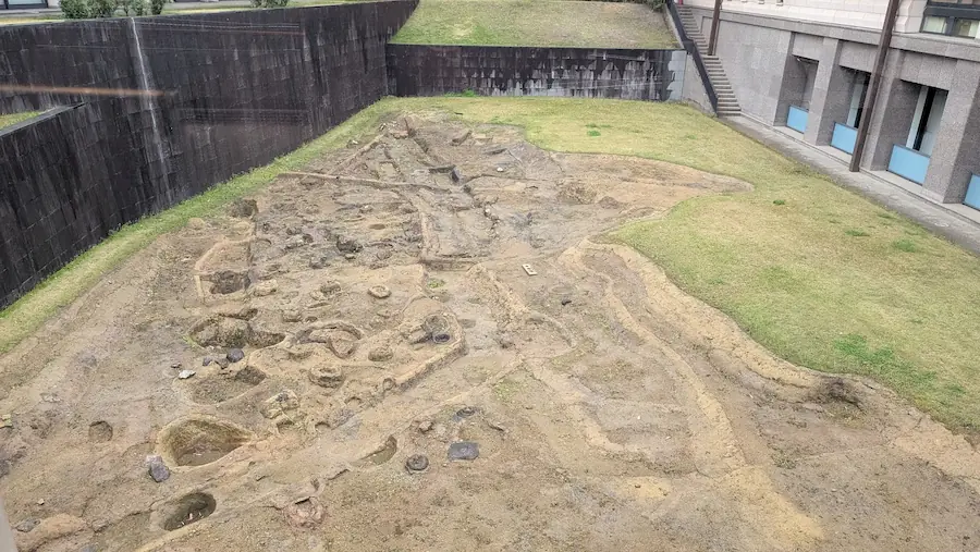 奈良県立万葉文化館の敷地内に保存されている飛鳥池遺跡。古代の工房跡の遺構が土に掘られた状態で露出し、周囲を近代的な建物に囲まれている様子。