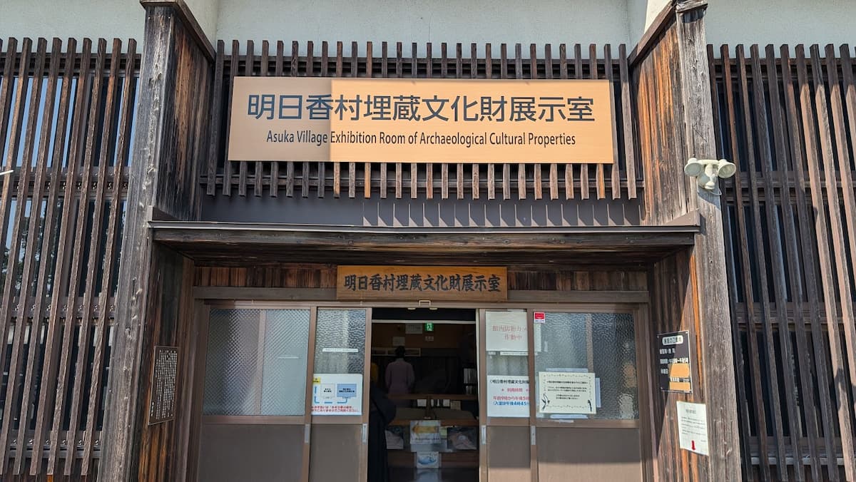 明日香村にある石神遺跡の敷地。左側に未舗装の散策路が伸び、右手には広大な草地(原っぱ)が広がっている。遠くにはなだらかな甘樫丘の山並みやのどかな民家が見える。